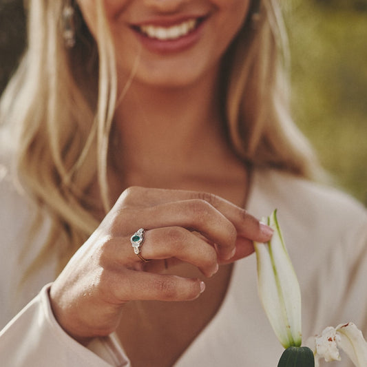 Mano tocando el pétalo de una flor y mostrando el precioso anillo Louise que destaca por su esmeralda.