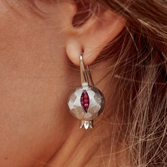 Primer plano de la oreja de una mujer con un pendiente de plata de ley con forma de granada. Tiene una pequeña abertura donde se ven unos rubíes simulando las semillas del fruto.