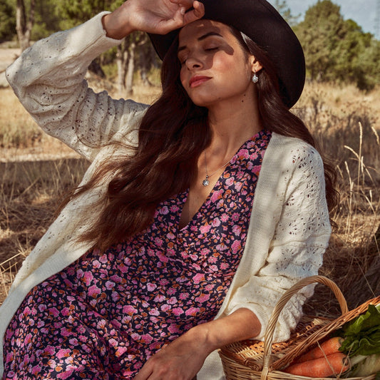 Modelo posando en el campo protegiéndose del sol con la mano y mostrando los pendientes Delphine. Se aprecia su forma de flor colgando de un pequeño arito de plata. 